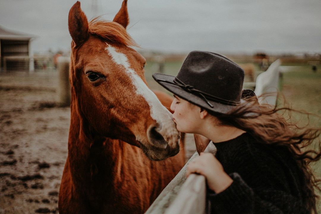 A woman in a felt hat leans in nose to nose with a chestnut horse at a fence